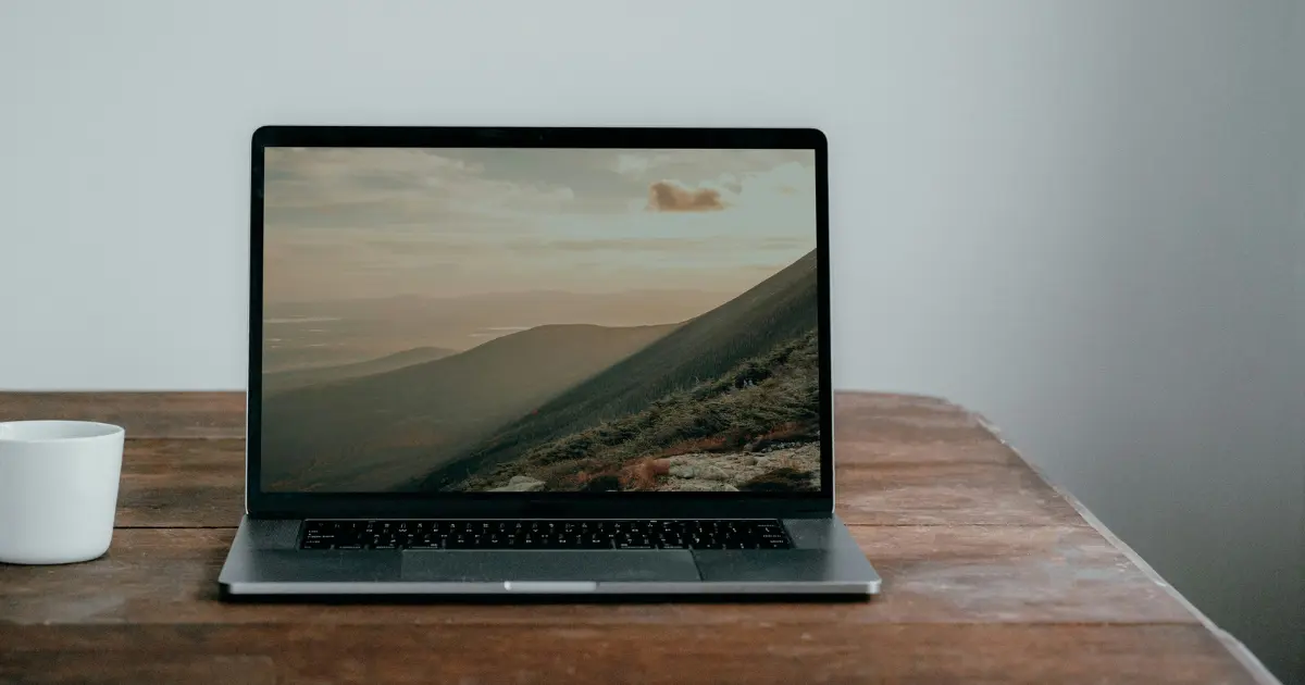 A laptop on a wooden table with a scenic background, alomgside a coffee cup.